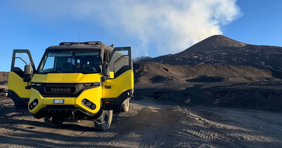 A yellow off-road vehicle parked on volcanic gravel on Etna North, the summit smoking behind