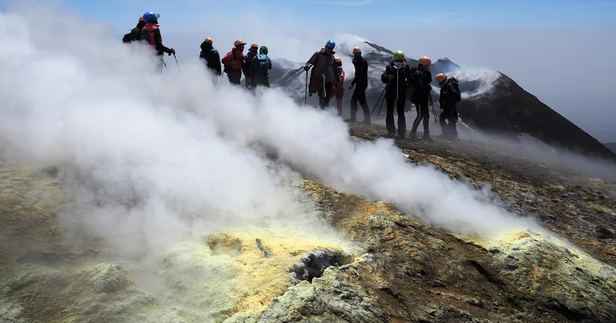 Hikers in helmets on the rim of a steaming summit crater of Etna, yellow sulphur on the rocks