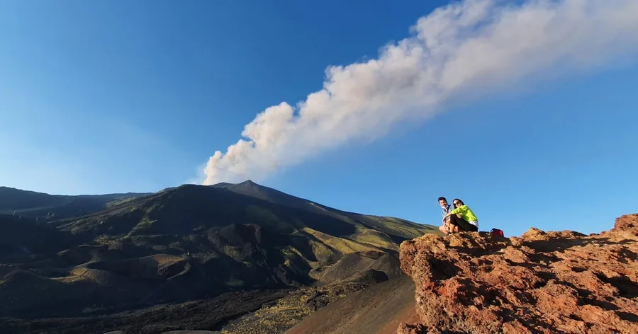A plume of white gas rising from Etna's summit over dark slopes, a hiker on a red rock outcrop