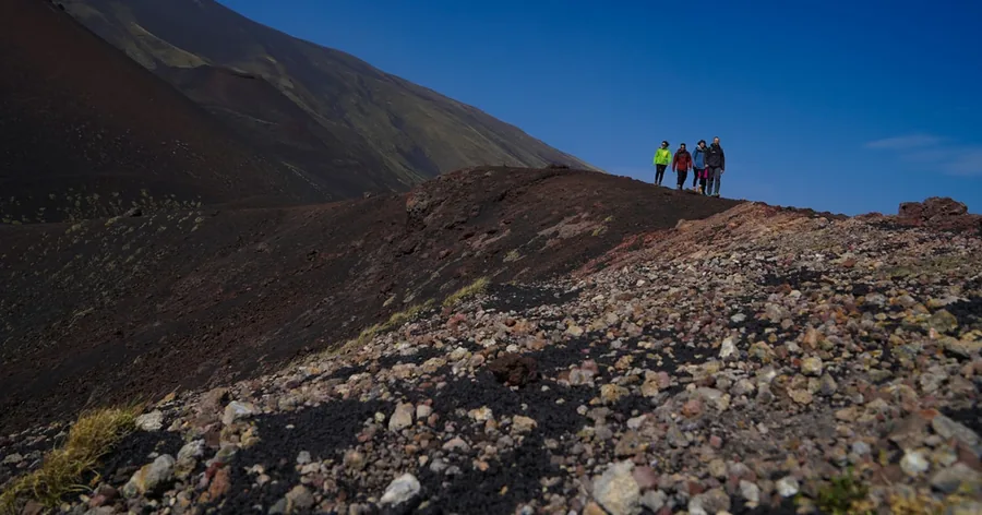 Hikers on a rust-coloured crater ridge high on Etna under a deep blue sky
