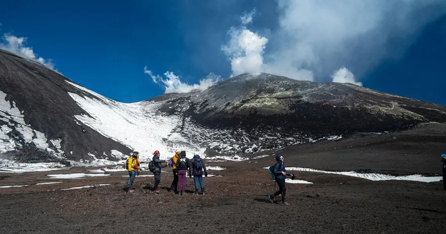 Hikers crossing a dark volcanic plain under the summit of Etna with late snow on its flanks