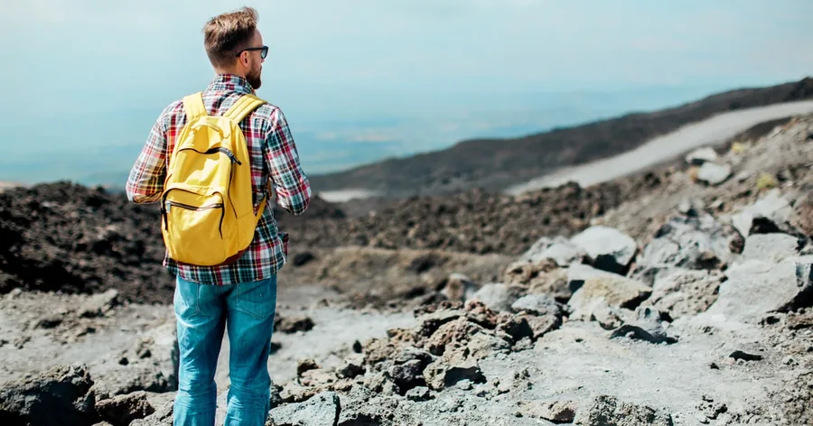 A hiker with a yellow backpack looking out over the grey lava fields of Etna towards the sea